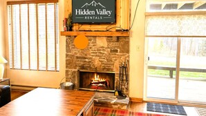A cozy living room with a stone fireplace, a television mounted on a wooden wall, and a large window with blinds. - Pool Access | Fireplace, Deck & A/C (Somerset)