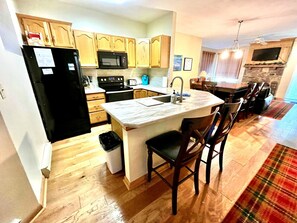 A kitchen with wooden cabinets, a black refrigerator, a microwave, a stove, and a dining area with chairs and a table. - Relaxing Mountain Condo with Fireplace & Deck (Somerset)