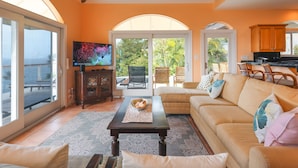 A spacious living room with a sofa, coffee table, and a television. There is a sliding glass door leading to an outdoor patio area with chairs and a table. - Oceanfront with Pool, Ocean Views, 5 Bedrooms! (St. Thomas)
