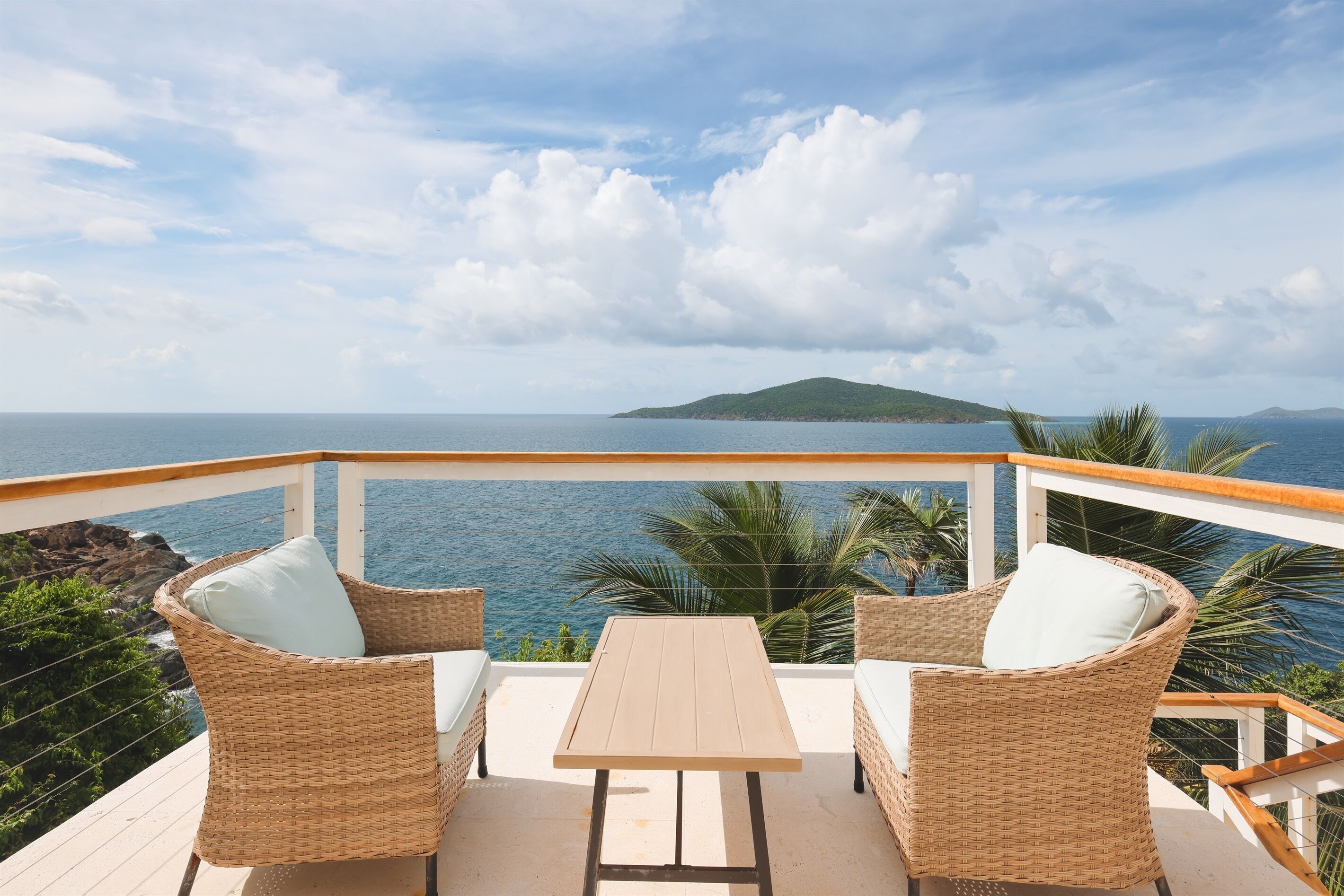 Un balcon agrémenté de meubles en osier et d’une table en bois, offrant une vue sur une étendue d’eau et une île lointaine.