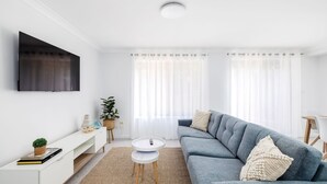 A modern living room with a grey sofa, a flat-screen TV mounted on the wall, a small round table, and a plant.