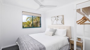 A bedroom with a bed, a ceiling fan, a window with a view of trees, a wall-mounted artwork, and a wooden wardrobe.