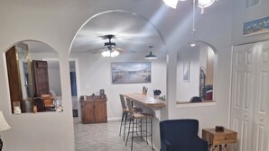 Een ruime woonkamer met een blauwe bank, een ronde houten salontafel en een eettafel met stoelen. Er is een plafondventilator, een schilderij aan de muur en een deur met een spiegel.