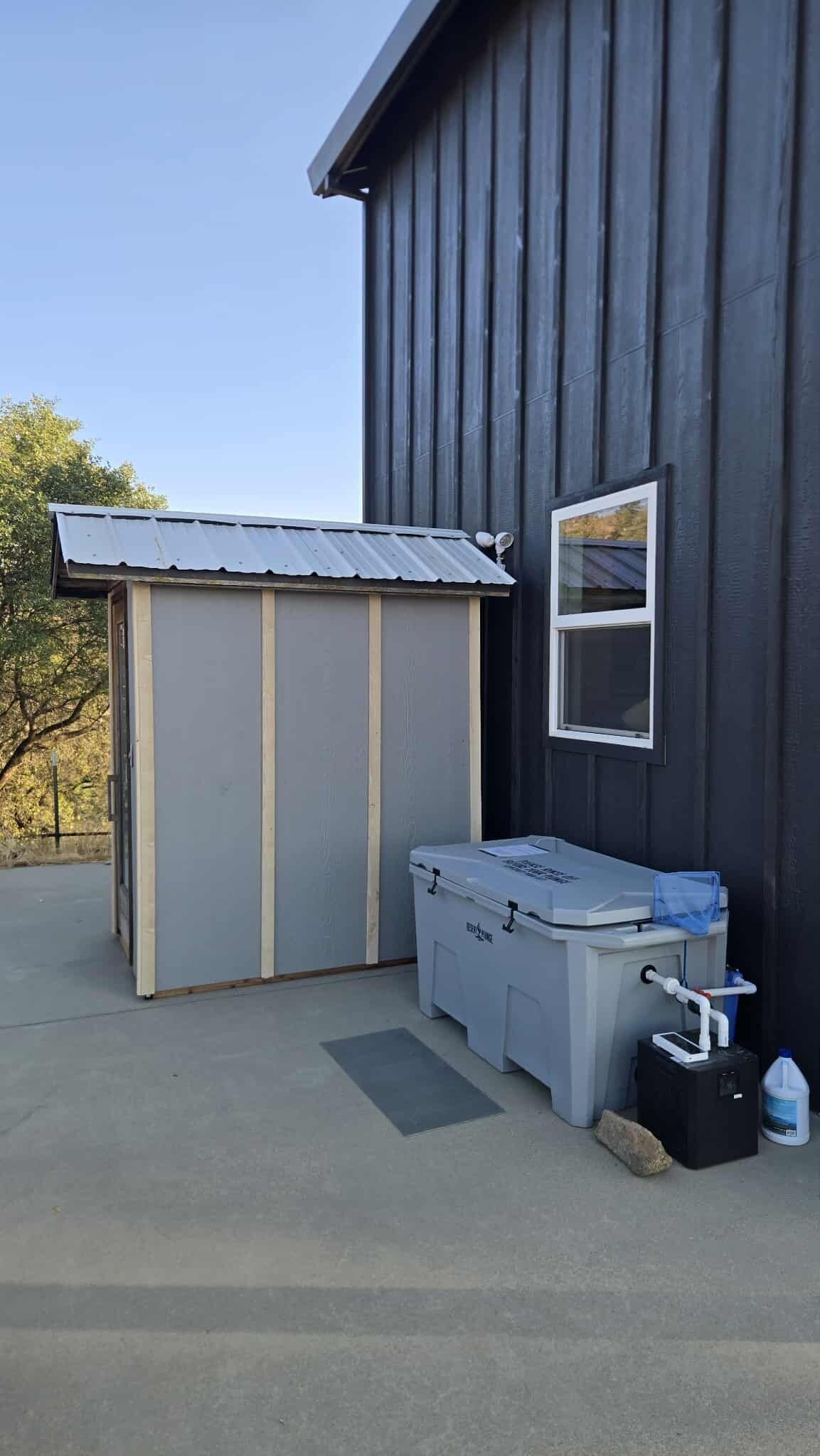 Un petit abri de jardin avec un toit en métal et une fenêtre, une boîte de rangement étiquetée « RECYCLAGE », et un générateur noir posé sur une surface en béton.