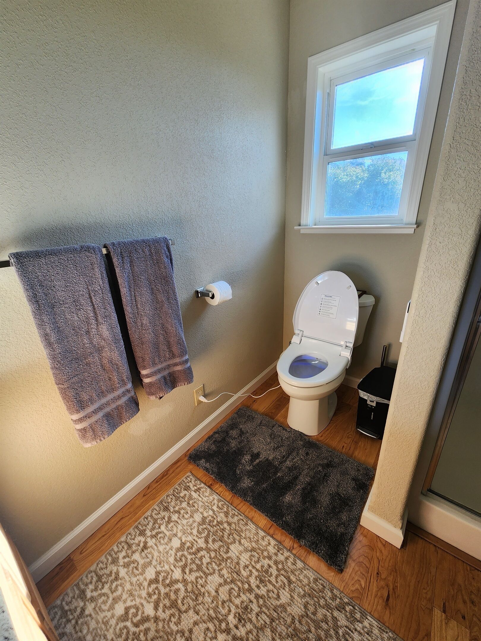 Un baño con dos toallas colgadas en la pared, un inodoro, una ventana y un cubo de basura.