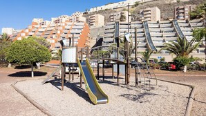 A playground with a slide and climbing structures, surrounded by a gravel area and palm trees. - Home2Book Fantastic 160m2 Terrace Flat & BBQ (Radazul)