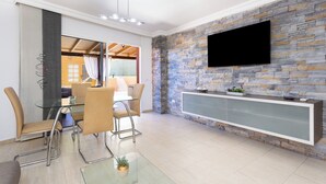 A modern dining area with a glass table and metal chairs, a stone accent wall, and a mounted TV. - Home2Book Fantastic 160m2 Terrace Flat & BBQ (Radazul)