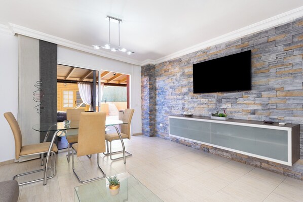 A modern dining area with a glass table and metal chairs, a stone accent wall, and a mounted TV.