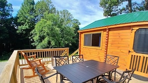 A wooden deck with a table and chairs, a metal trash can, and a cabin with a green roof.