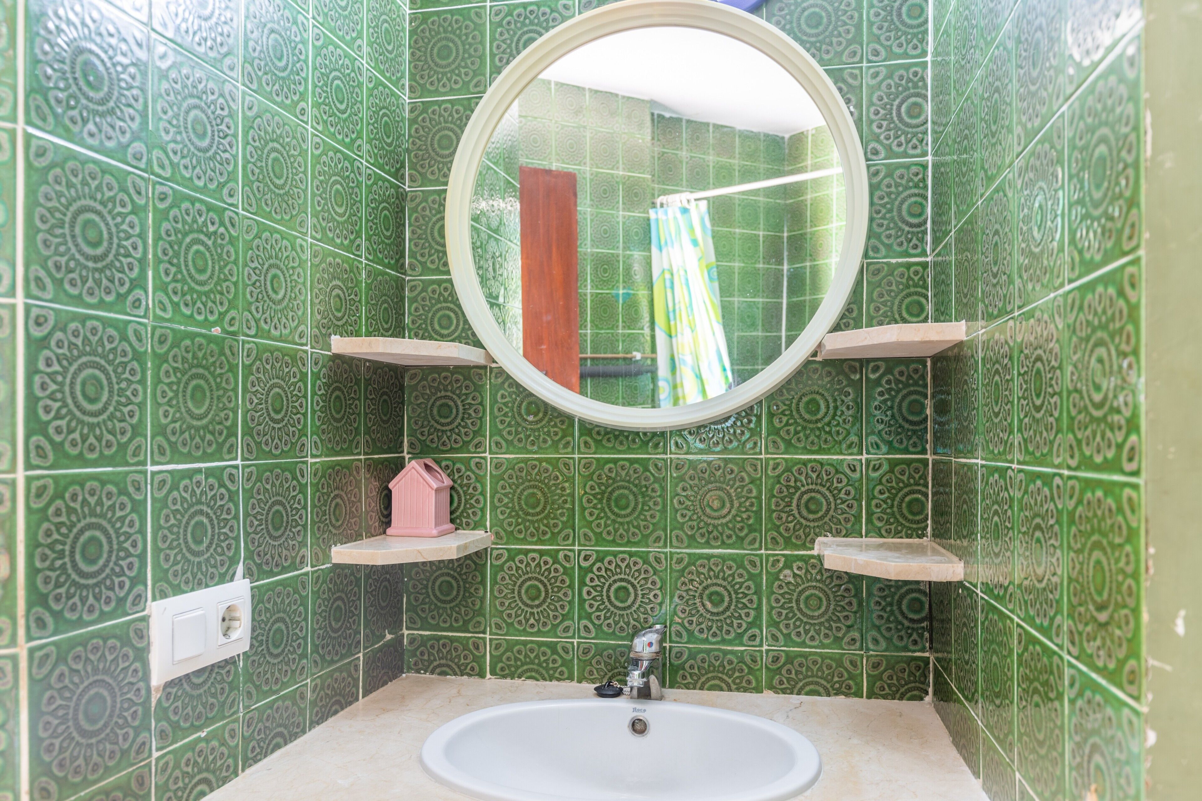 A bathroom with green patterned tiles, a round mirror, a sink, and two shelves.