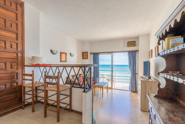 Um quarto com porta de madeira, área de jantar com cadeiras, mesa e vista para o mar por uma porta de vidro.
