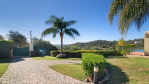 A paved driveway leading to a gated entrance, a palm tree, and a view of a hilly landscape. - Holiday Home "Casa no Campo" with Sea View, Shared Pool and Wi-Fi (Loulé)
