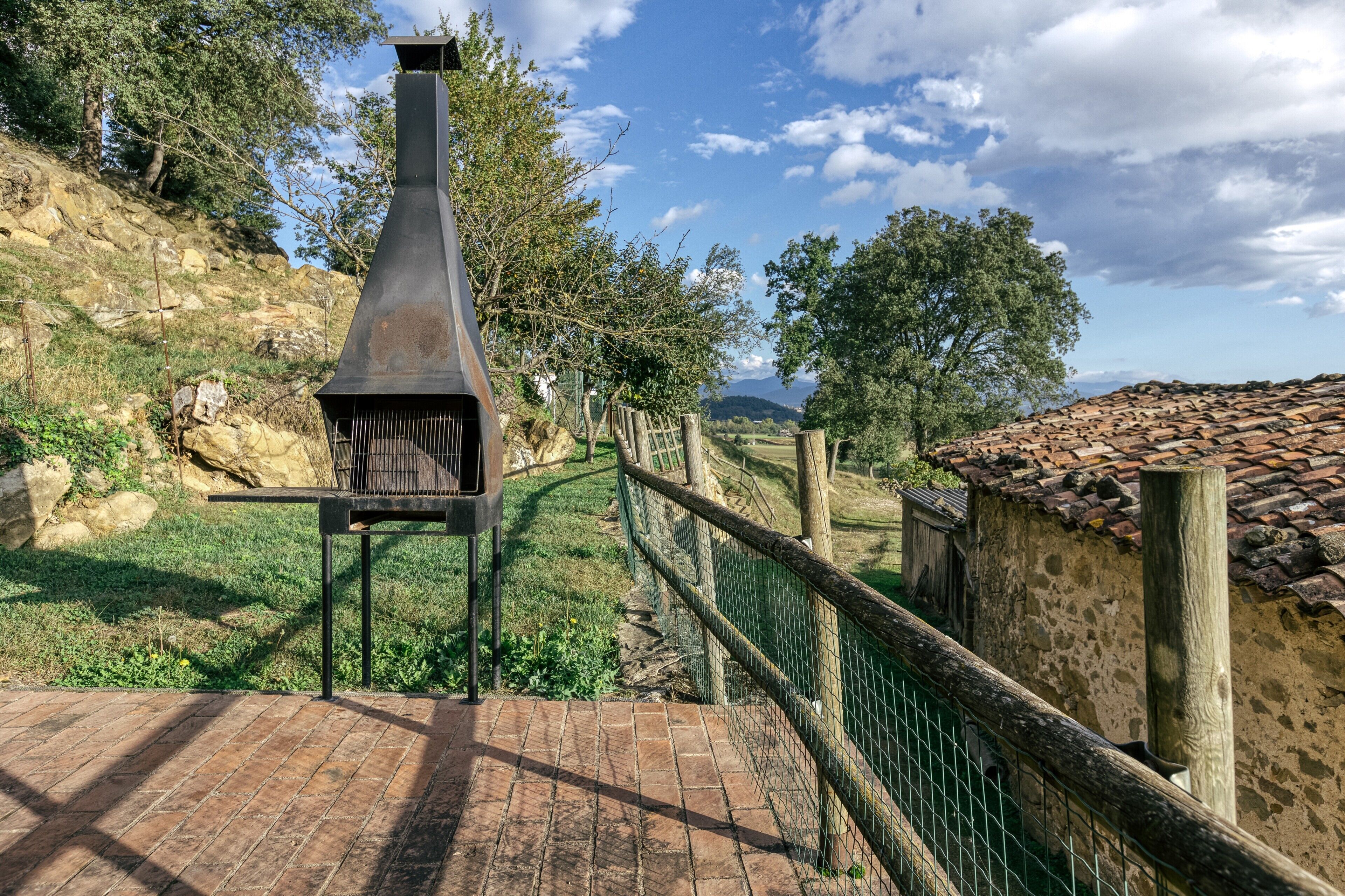 A rustic outdoor grill area with a brick pathway, a wooden fence, and a stone wall.
