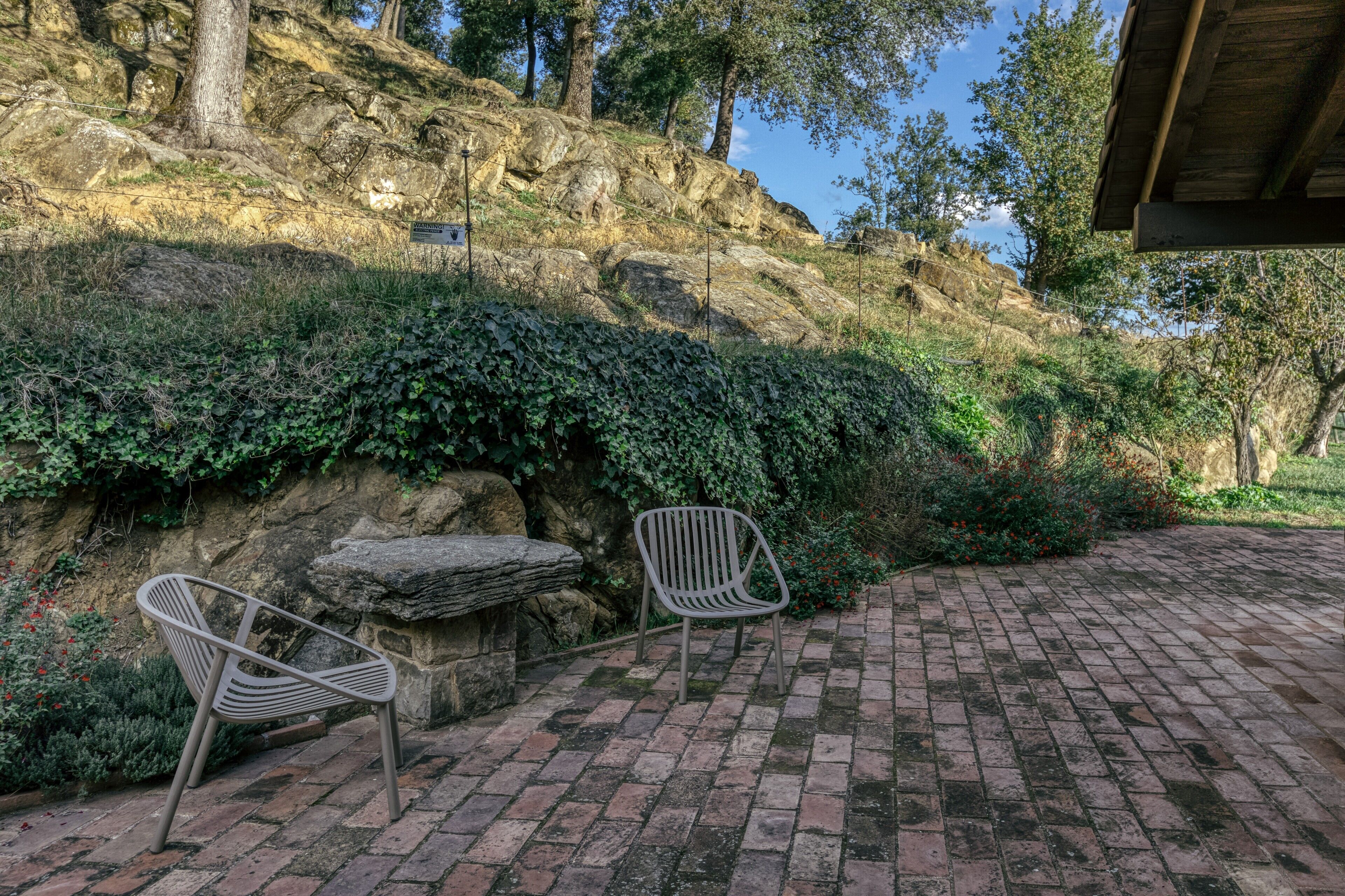 Un patio empedrado con dos sillas de metal, un banco de piedra y un paisaje de colinas rocosas al fondo.