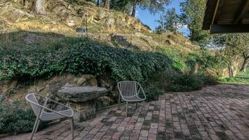 Un patio empedrado con dos sillas de metal, un banco de piedra y un paisaje de colinas rocosas al fondo.