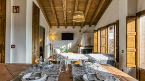 Un coin repas avec une longue table dressée pour deux personnes, des poutres en bois au plafond, une cheminée et un téléviseur fixé au mur.