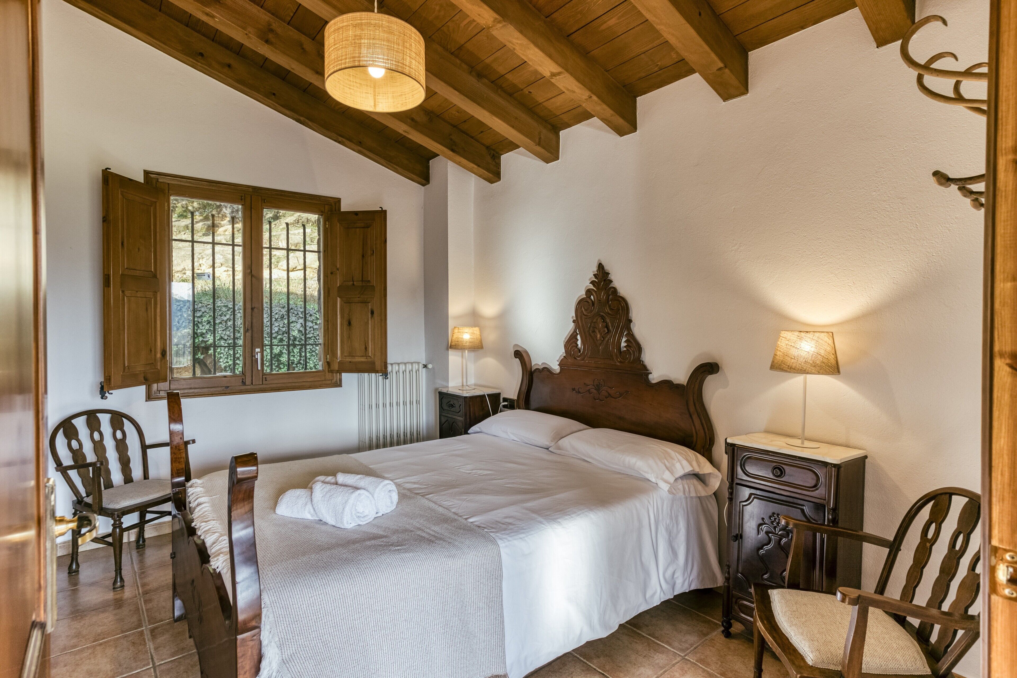 Un dormitorio con una cama de madera, dos sillas, una mesita de noche, una lámpara y una ventana con contraventanas de madera.