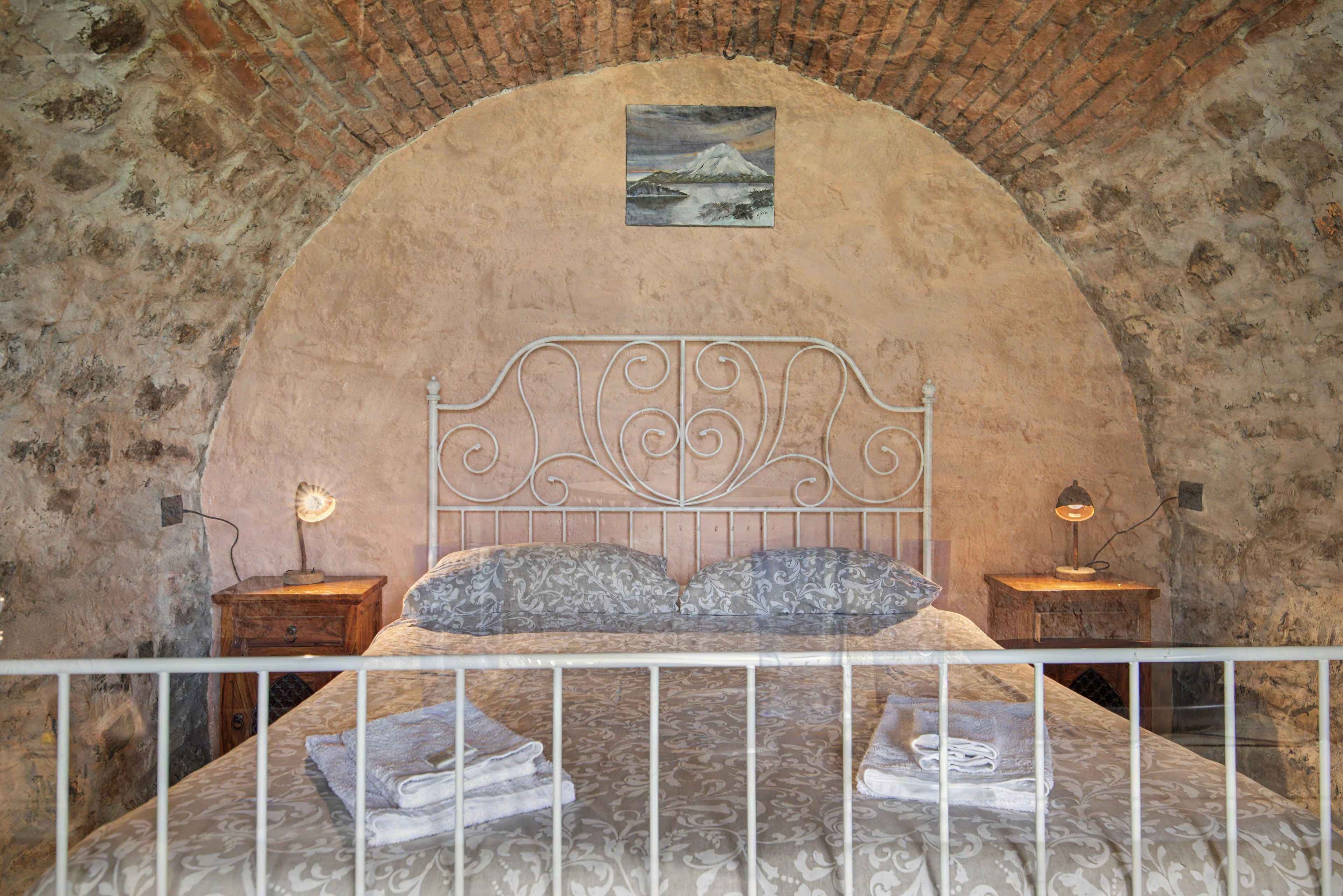 A bedroom with a stone wall, a bed with a metal headboard, two bedside tables with lamps, and a painting above the bed.
