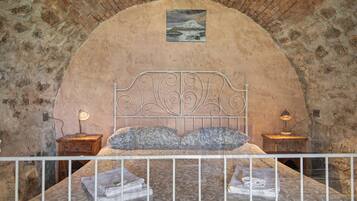 A bedroom with a stone wall, a bed with a metal headboard, two bedside tables with lamps, and a painting above the bed.