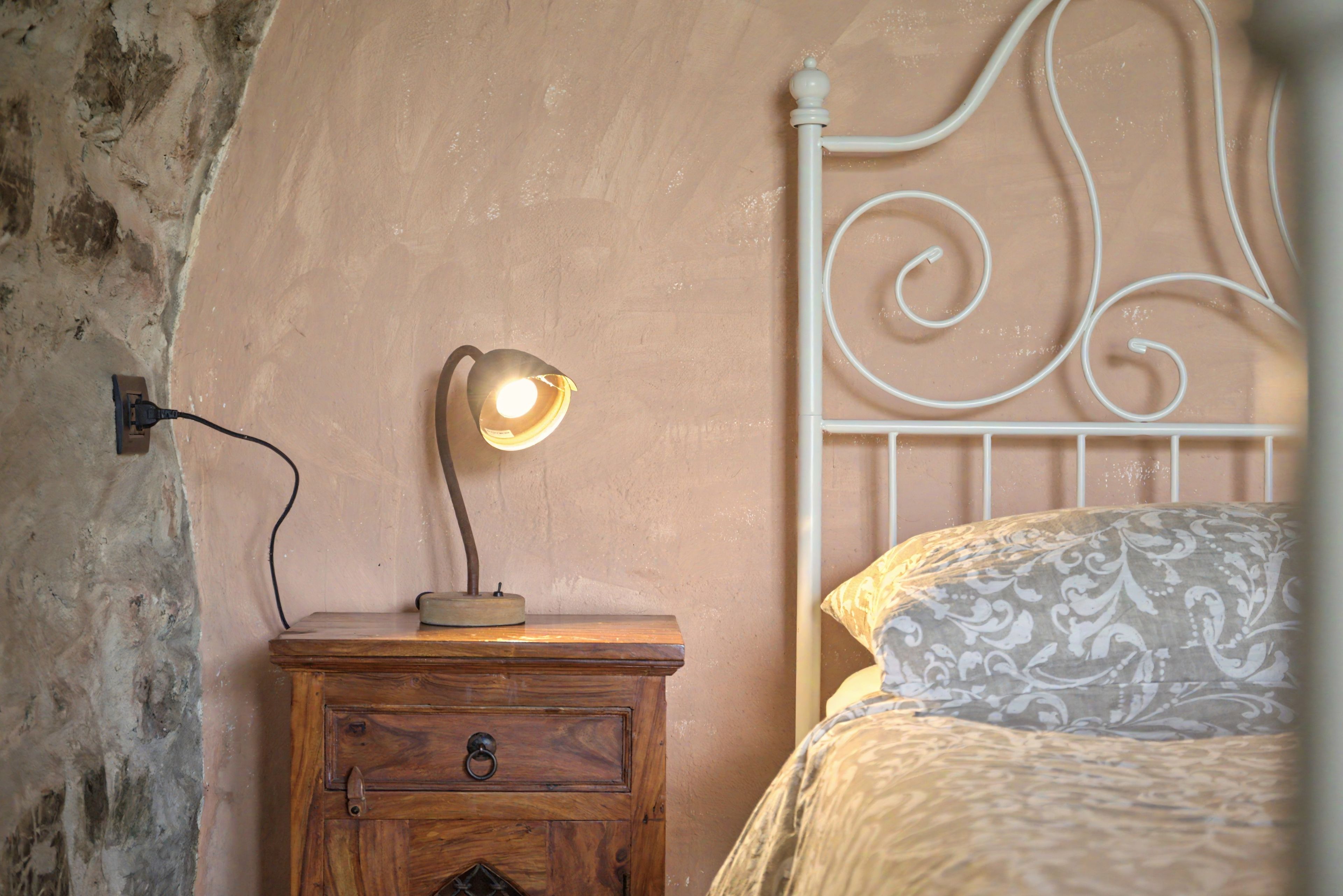 A bedroom with a wooden nightstand, a lamp, a bed with a patterned pillow, and a white metal headboard.