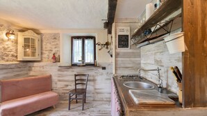 Una cocina rústica con pared de piedra, vigas de madera, un sofá rosa, una mesa de comedor y un fregadero.