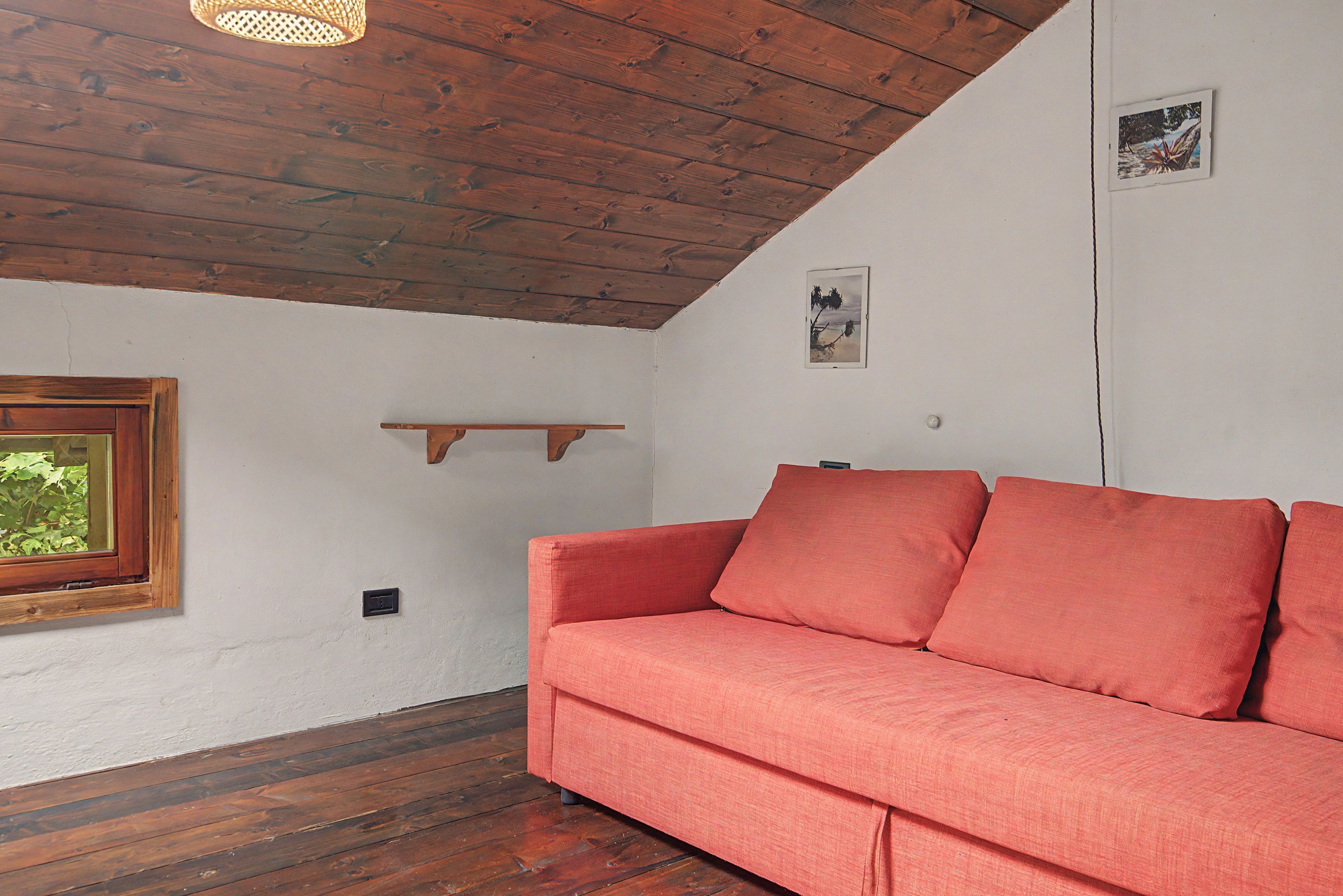 Een kamer met een houten plafond, een rode bank, een raam met uitzicht op groen en een klein schap aan de muur.