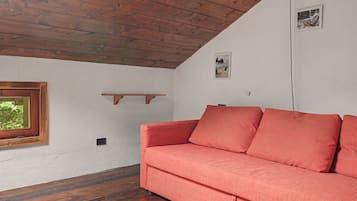 A room with a wooden ceiling, a red sofa, a window with a view of greenery, and a small shelf on the wall.