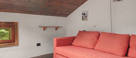 A room with a wooden ceiling, a red sofa, a window with a view of greenery, and a small shelf on the wall.