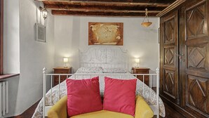 Une chambre avec un plafond en bois, un lit recouvert d’une couette blanche à motifs, deux oreillers rouges, une chaise jaune et une armoire en bois.