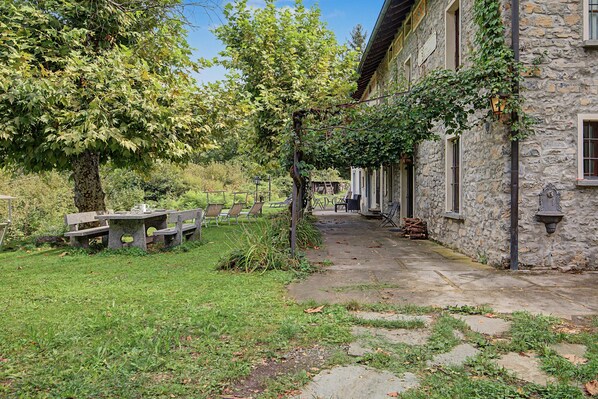 A stone building with a covered outdoor seating area, a paved pathway, and a tree with green leaves.
