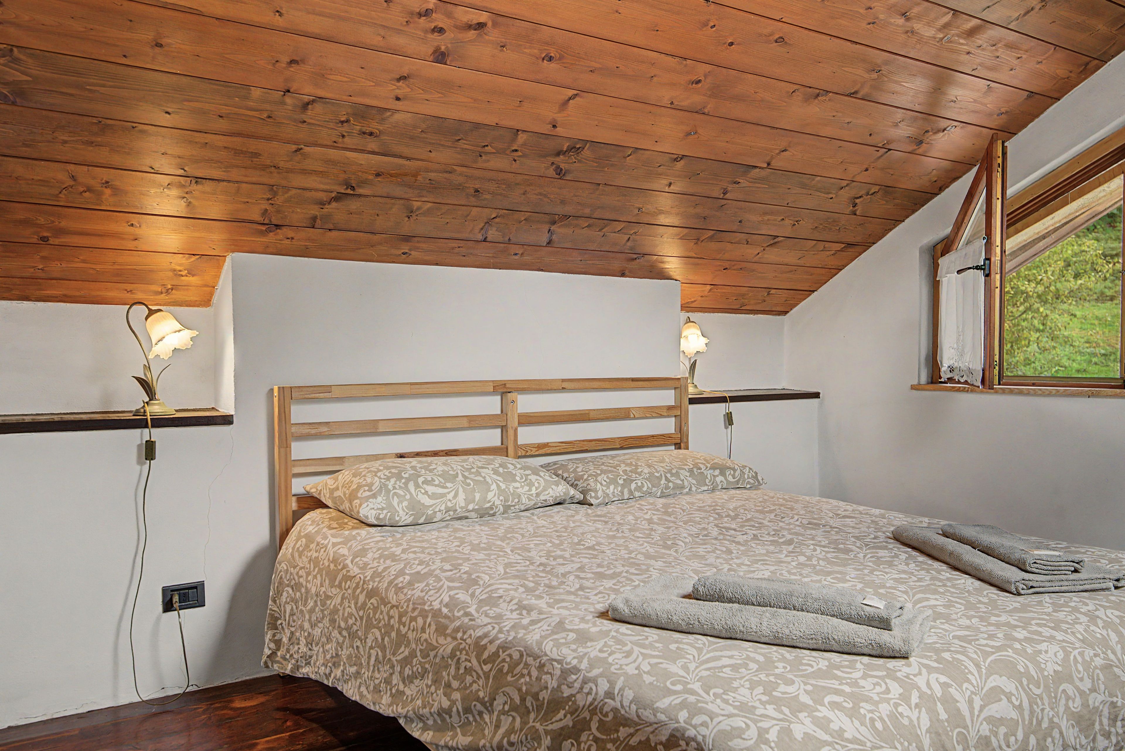 A bedroom with a wooden headboard, two bedside lamps, a bed with patterned bedding, and a window with a view of greenery.