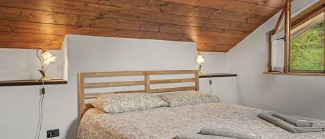 Ein Schlafzimmer mit einem hölzernen Kopfteil, zwei Nachttischlampen, einem Bett mit gemusterten Bettwaren und einem Fenster mit Blick auf Grünflächen.