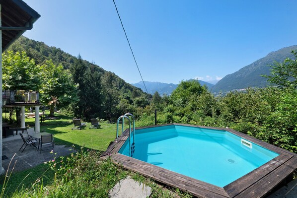 Una piscina rettangolare con un solarium in legno, circondata da un rigoglioso giardino e uno sfondo montuoso.