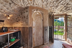 A rustic interior with a stone ceiling, a wooden door, a view of greenery outside, and a shelf with kitchen items.