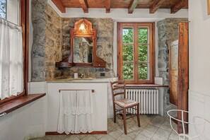 A rustic bathroom with stone walls, a wooden vanity, a mirror, a radiator, and a window with a view of greenery.