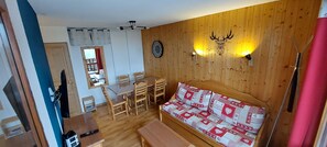 A room with a wooden wall, a sofa with patterned cushions, a dining table with chairs, a clock, and a mounted television. - Apartment "Valmeinier 1800" with Mountain View & Shared Pool (Valmeinier)