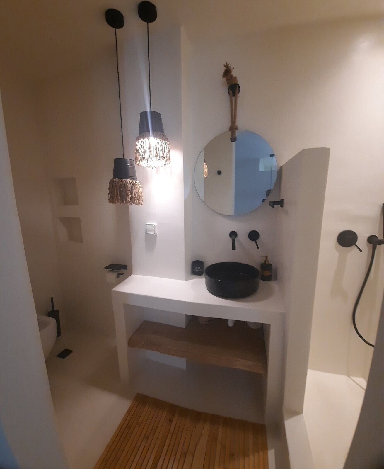 Un baño con lavamanos blanco, espejo redondo, lámpara colgante y zona de ducha.
