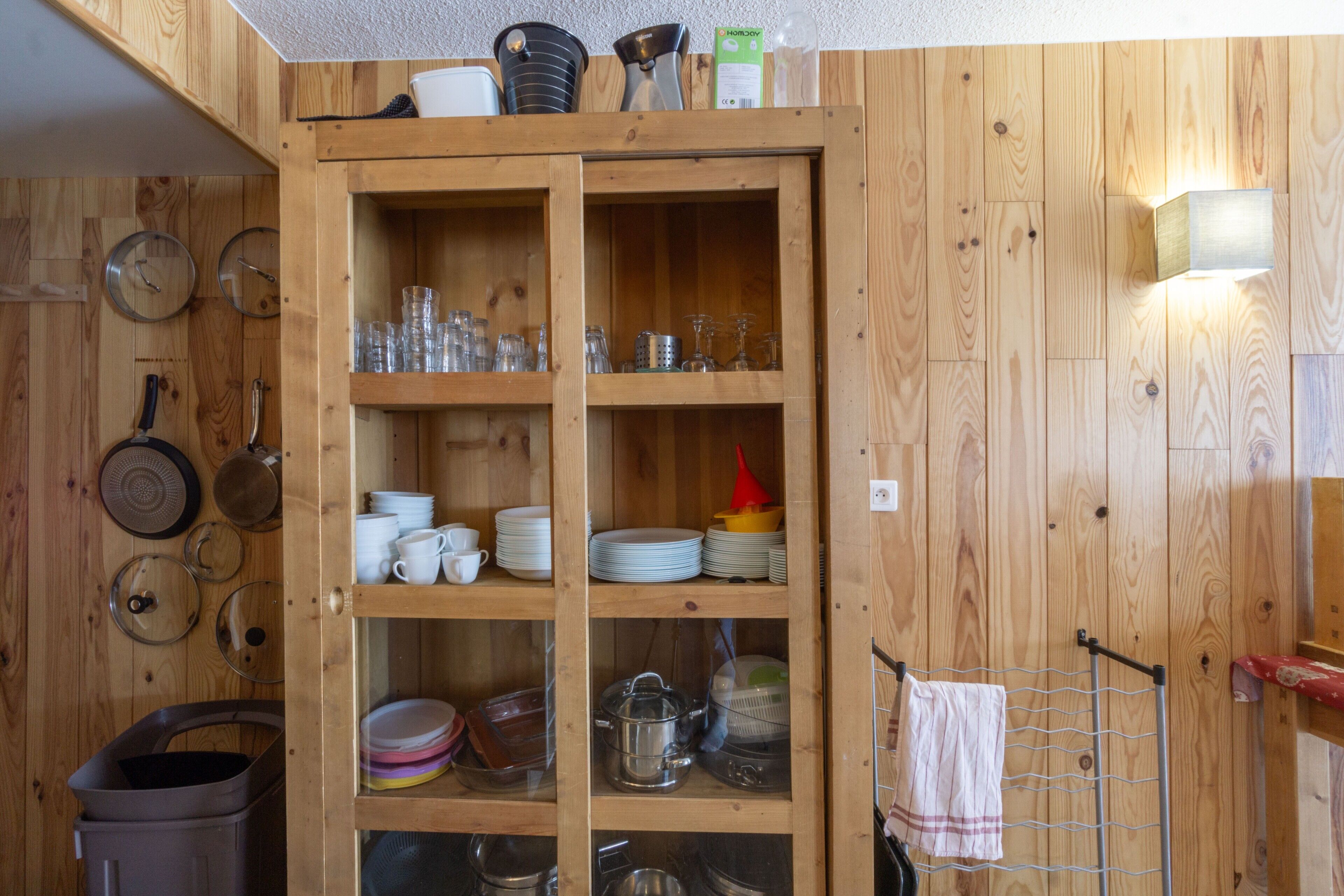 Un meuble en bois doté d’étagères où sont rangés des ustensiles de cuisine, un porte-serviettes suspendu et un luminaire fixé au mur.