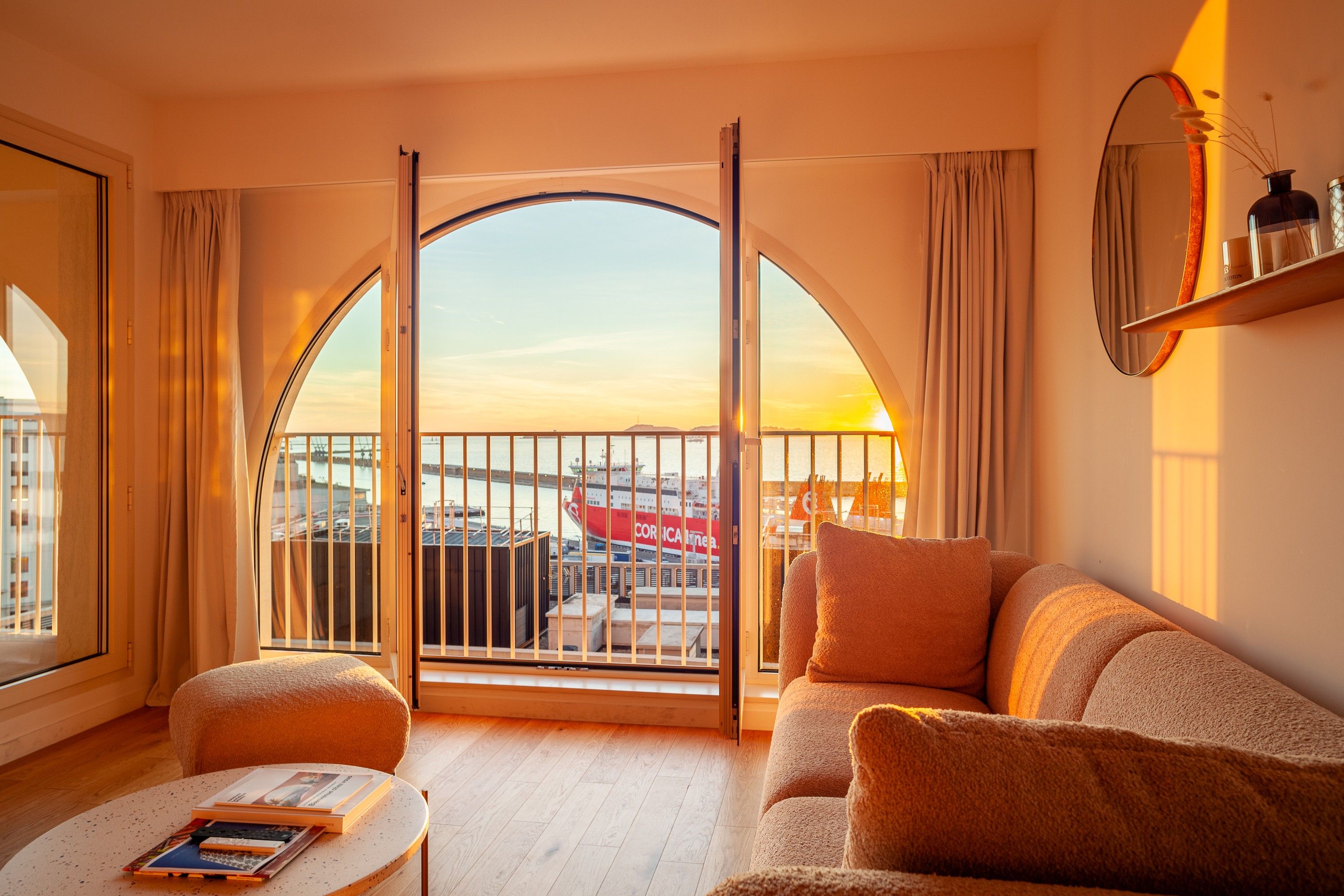 A hotel room with a large arched window, a sofa, a small table with books, and a view of a ship at the dock.