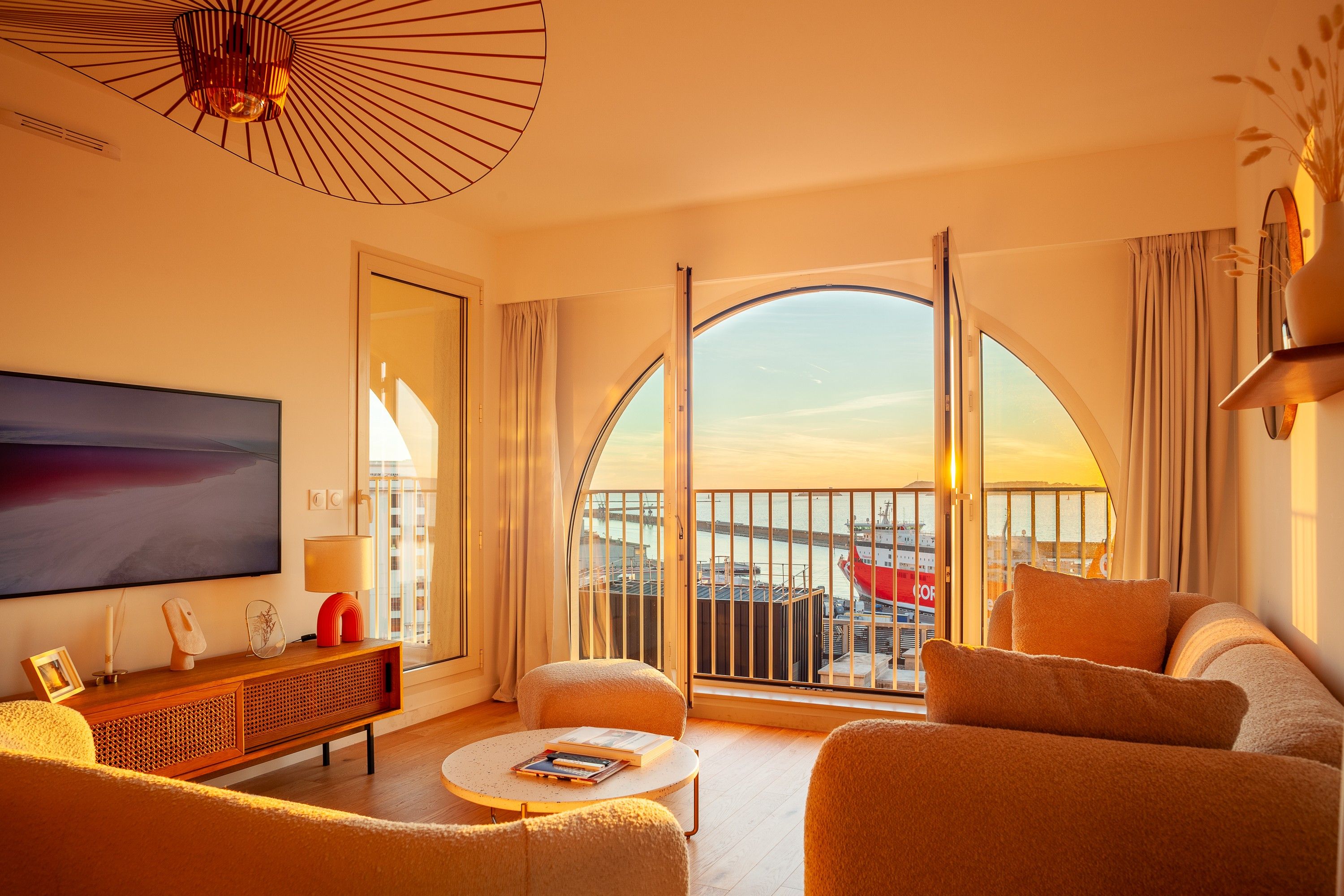 A modern living room with a large TV, comfortable seating, a coffee table, and a view of a port with ships.