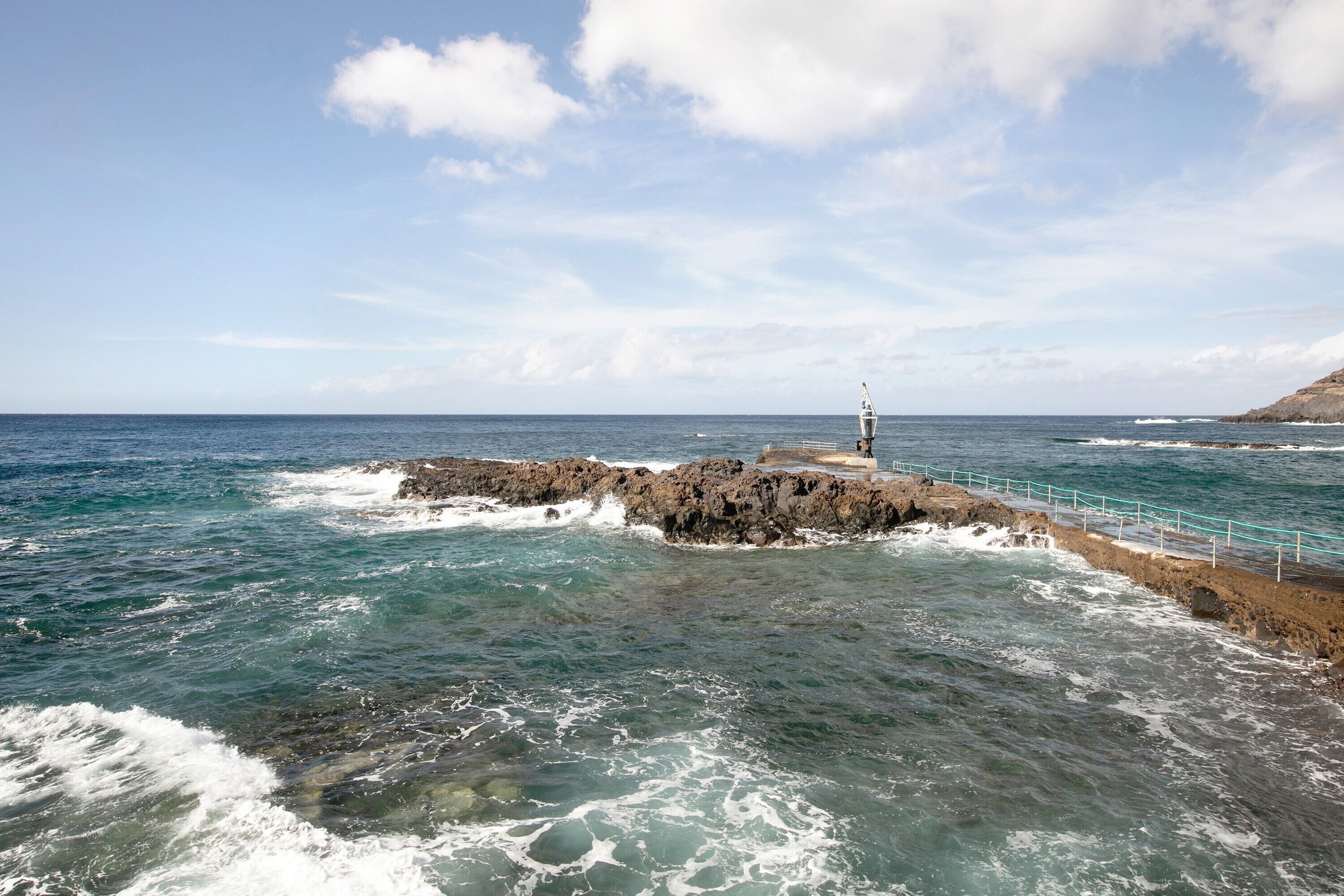 Paisaje costero con un rompeolas rocoso, un faro y cielo despejado.