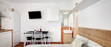 Un dormitorio con cama, mesita de noche, televisor, una pequeña mesa con sillas, cocina equipada con refrigerador y baño con espejo y lavamanos.