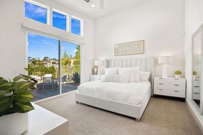 Un dormitorio moderno con una cama grande, una mesita de noche, una lámpara y un espejo. Hay una puerta corrediza de vidrio que da a un balcón con plantas y vistas al exterior.