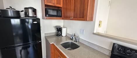 Cocina compacta con armarios de madera, un refrigerador negro, microondas, cafetera y pileta.