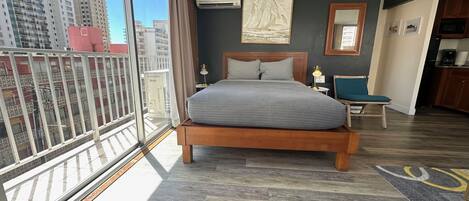 Habitación de hotel con una cama grande, un escritorio pequeño, una silla, un balcón con barandilla y vistas a edificios altos.