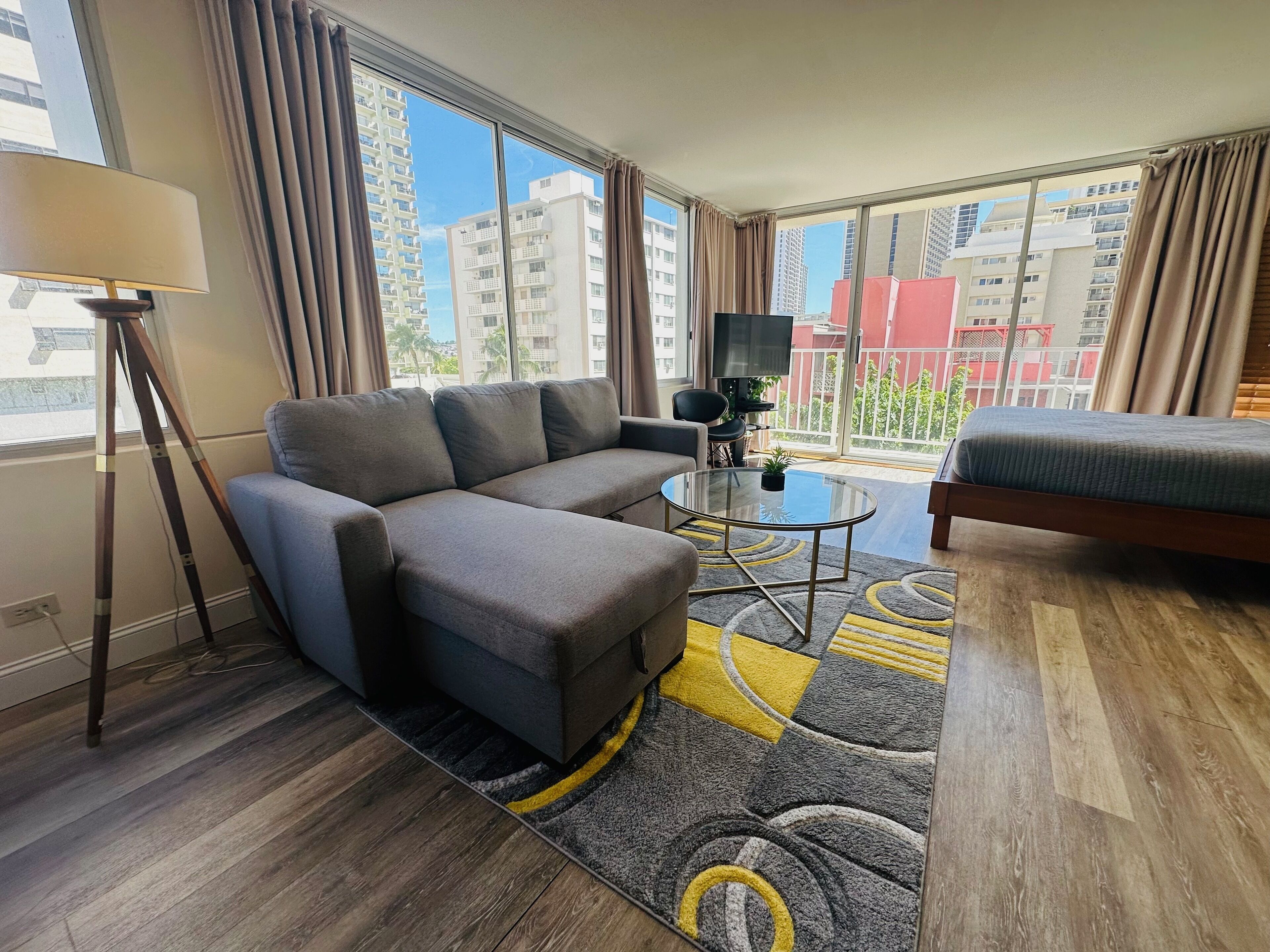 Un salon moderne avec un canapé d’angle, une petite table basse ronde, un téléviseur et un balcon offrant une vue sur les immeubles de grande hauteur.