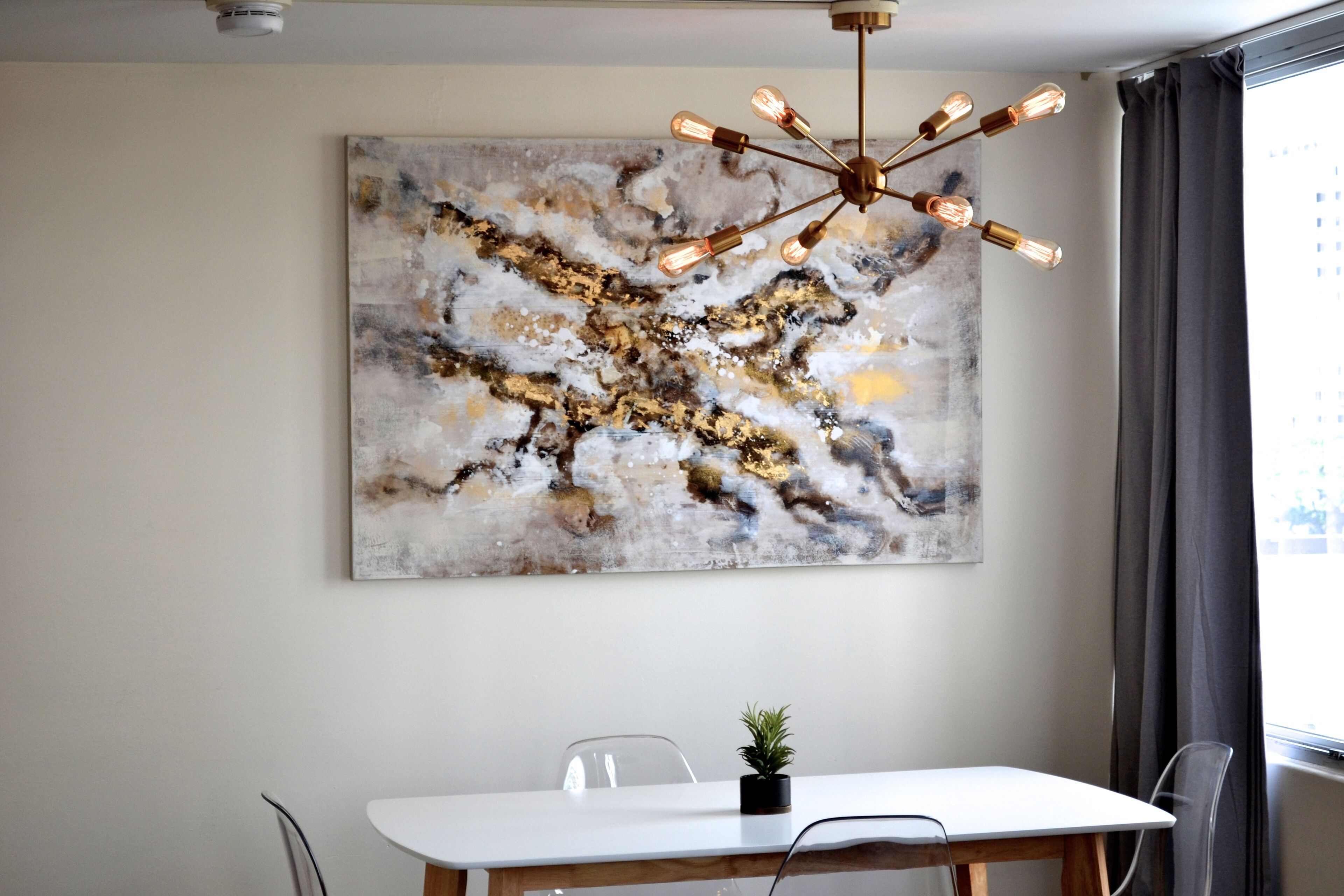 A dining area with a white table, clear chairs, a modern chandelier, and a large abstract painting on the wall.
