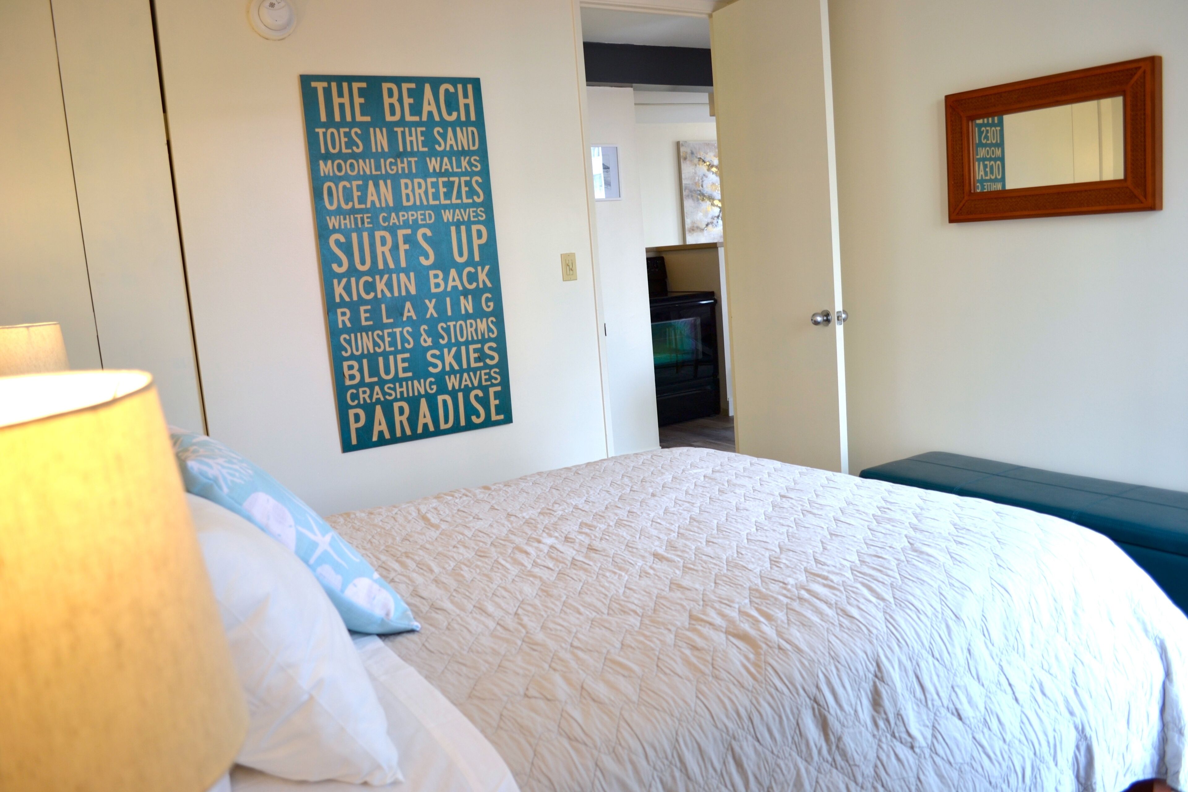 A bedroom with a bed, a wall with a beach-themed poster, a lamp, and a mirror.