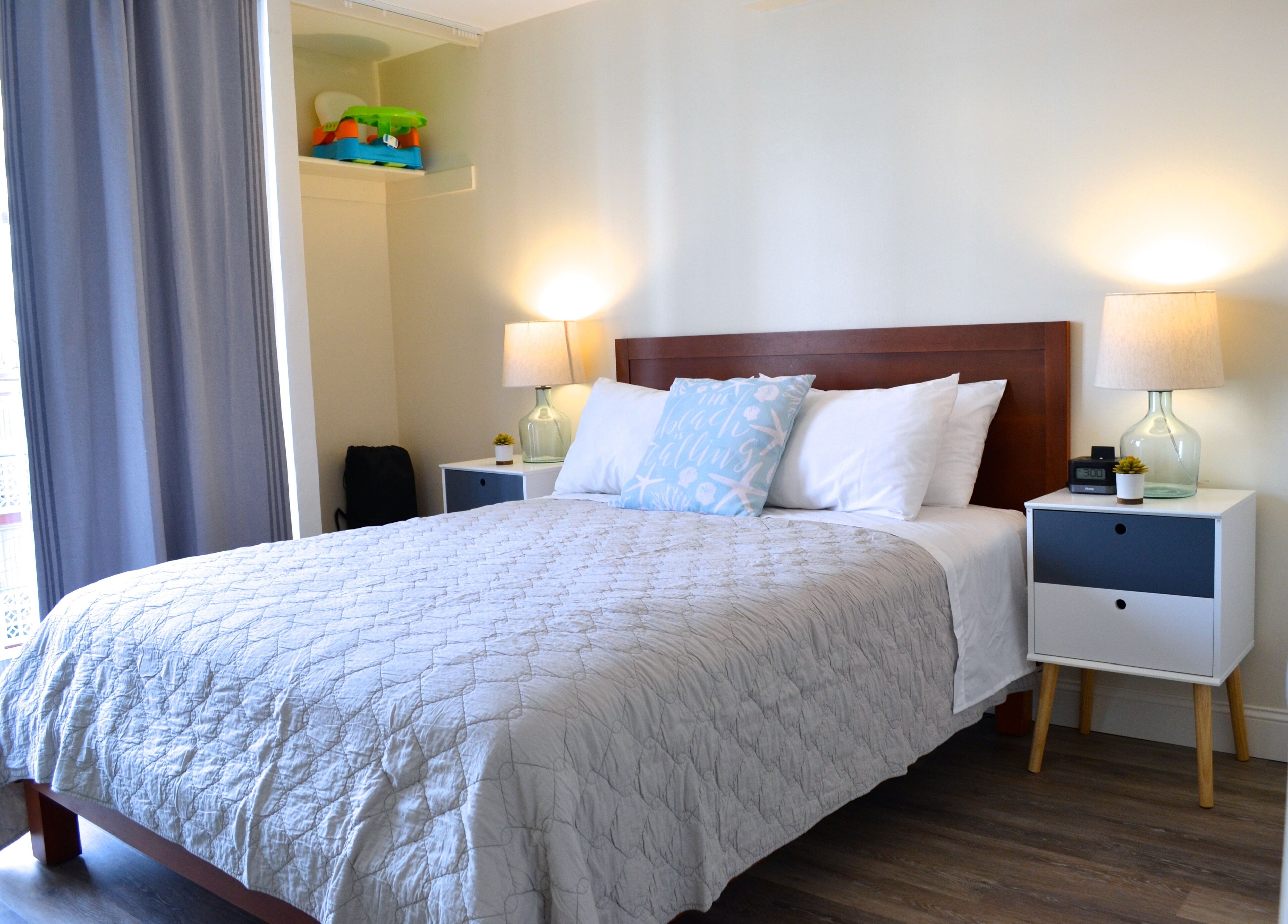 A neatly made bed with a quilted white cover, two bedside tables with lamps, a wooden headboard, and a small shelf above the bed.