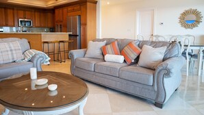 Una sala de estar moderna con un sofá gris, una mesa de centro de vidrio y una zona de cocina al fondo.